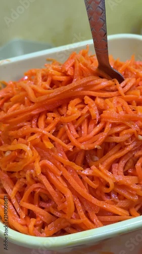 Close-up Korean carrot salad container, bright orange shredded carrots tossed with sesame and garlic, glossy oil sheen, spicy
