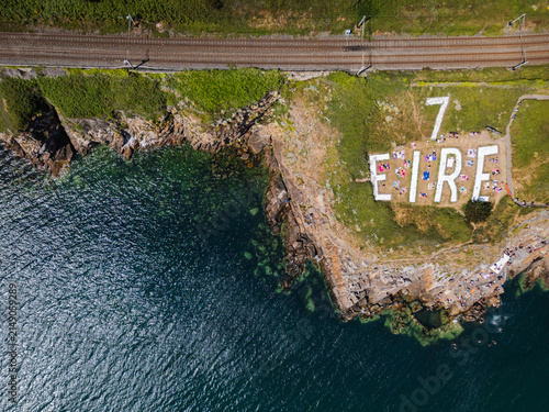 Aerial view of the historic EIRE 7 neutrality sign on the rocky coastline of Killiney Beach with people relaxing on the grass near the railway line in Dublin, County Dublin, Ireland.