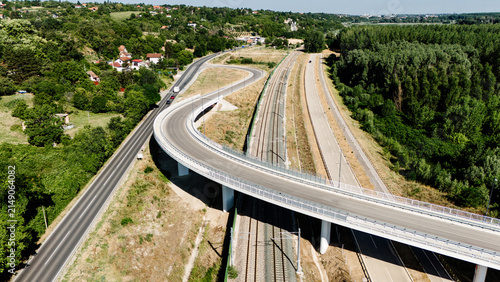 Aerial view of a modern highway viaduct curving over a railway line, surrounded by lush green trees and rural landscape in Sremski Karlovci, Vojvodina, Serbia.