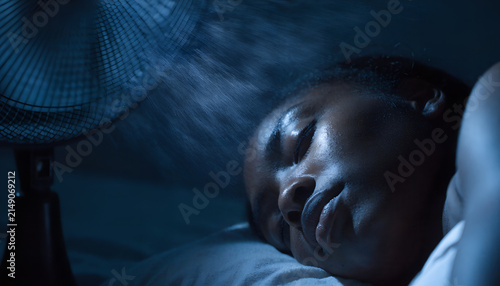 Young Black woman sleeping peacefully in a comfortable bed during a hot summer night, refreshed by the gentle breeze of a modern pedestal fan in a cozy dark bedroom.