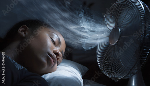 Young Black woman sleeping peacefully in a comfortable bed during a hot summer night, refreshed by the gentle breeze of a modern pedestal fan in a cozy dark bedroom.