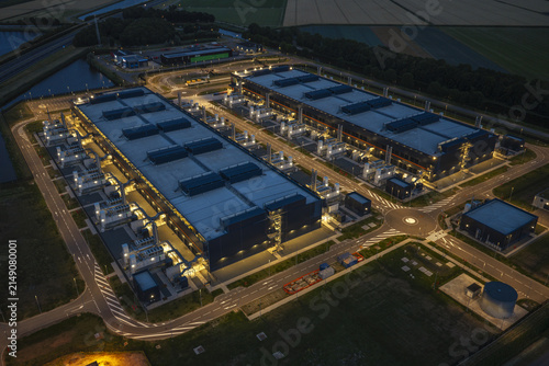 Aerial view of a large hyperscale data center illuminated at night, featuring expansive industrial buildings, cooling infrastructure, and surrounding road networks in the Netherlands.