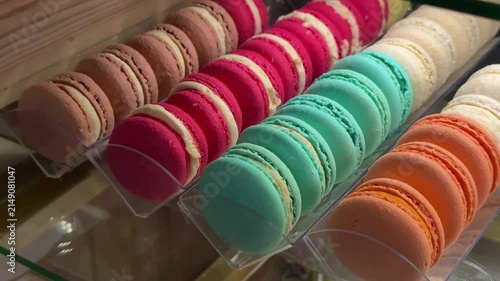 Colorful macarons coffee shop window close-up, rows of teal pink orange and brown shells arranged on clear tray inside warm lit