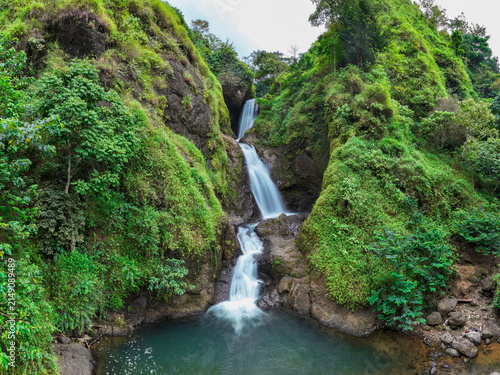 Aerial view of Curug Jagapati waterfall cascading down a lush, green, moss-covered rocky cliff into a clear pool in Cisompet, West Java, Indonesia.