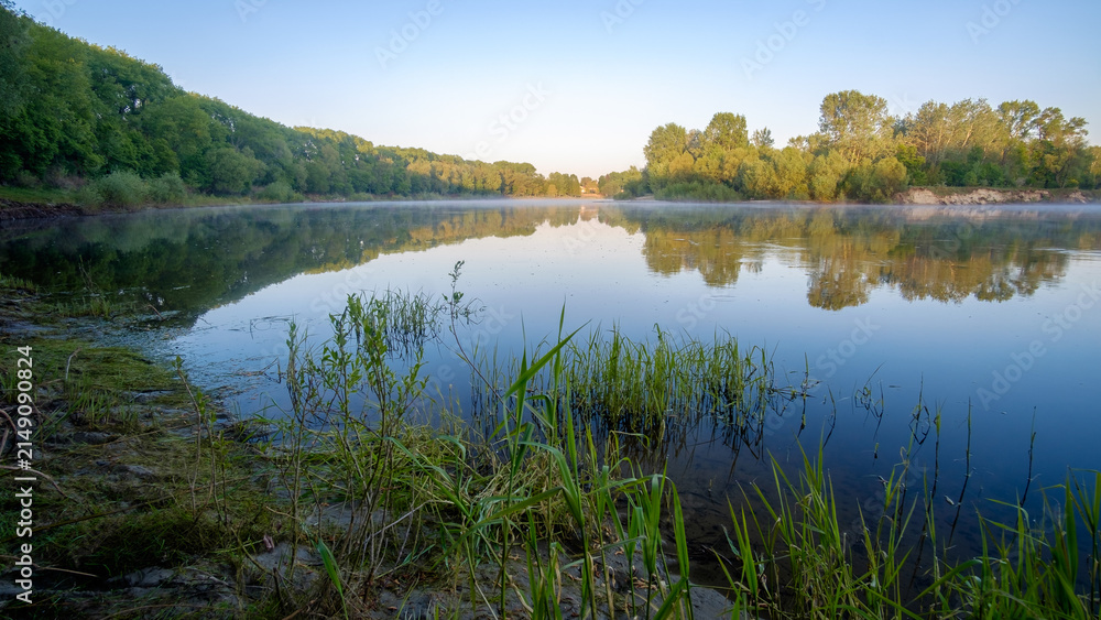 Obraz premium A calm river reflects lush green trees and a clear blue sky on a tranquil, bright and sunny morning.
