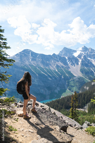 View of a woman hiking on a rocky trail overlooking the turquoise waters of Lake Louise surrounded by snow-capped mountains and pine forests in Lake Louise, Alberta, Canada.