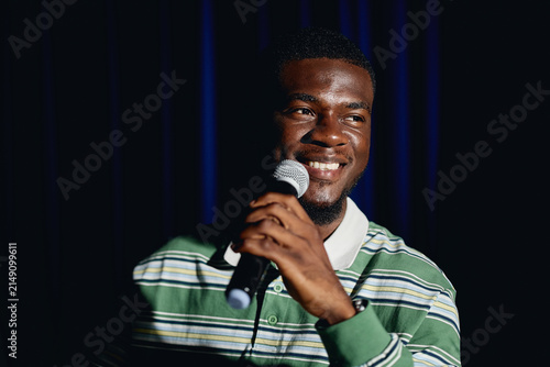 African American young adult male student smiling while holding microphone on theater stage. Image represents public speaking and performing arts education, medium shot