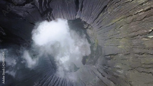 Aerial view of the volcanic crater of Mount Bromo with swirling white smoke rising from the deep, rugged, and textured dark grey walls, Tosari, East Java, Indonesia.