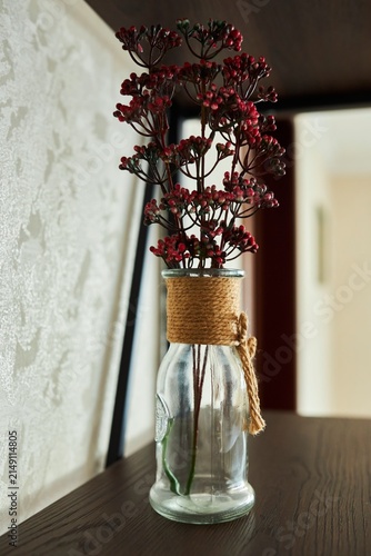 Red berries decorate a clear glass vase. The neck is wrapped with twine. The vase stands on a dark wood surface.