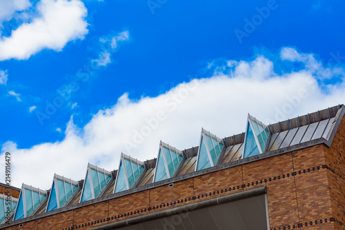 Brown brick wall with a distinctive triangular glass panel roof under a bright sky filled with soft, scattered clouds.