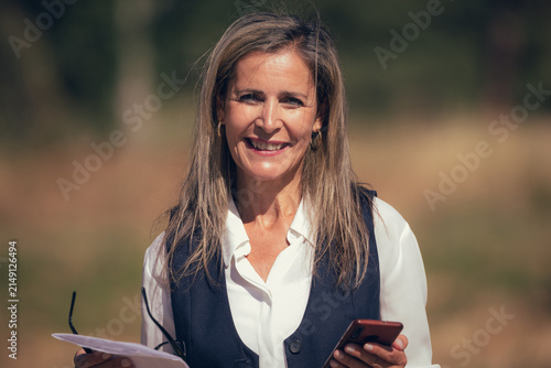 Smiling confident Caucasian businesswoman with long brown hair standing outdoors, holding papers and glasses, looking at camera with genuine happy expression in natural sunlight