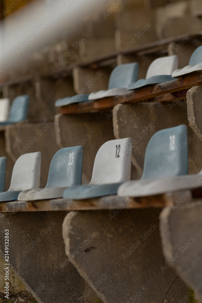 Fototapeta premium A row of numbered molded plastic seats fixed to aged concrete bleachers in an outdoor stadium. Close framing shows seat numbers, muted blue and gray seat