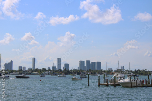 Scenic view of a coastal marina with moored motorboats and yachts along wooden docks, overlooking a distant modern urban city skyline under a blue sky with scattered clouds.