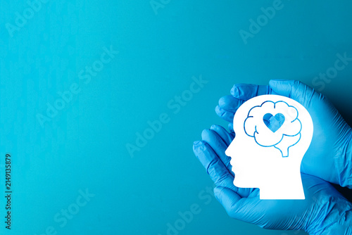 Mental health care and psychology concept. Doctor hands in blue medical gloves holding a human head silhouette with brain and heart on a solid blue background with copy space.