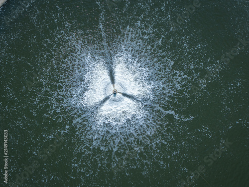 Aerial view of a water aerator creating a star-shaped pattern of bubbles and ripples on the surface of a dark pond in Poland.