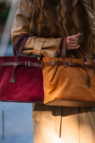 Close view of beige leather tote in woman hand with long wavy brown hair, ringed fingers, stitched seams, buckles, and tan straps during urban daytime fashion moment