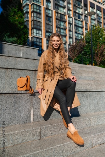 Smiling young woman seated on city steps in beige trench coat with black pants, brown boots, and tan handbag beside her in bright urban outdoor setting, with woman, smiling, female, person detail