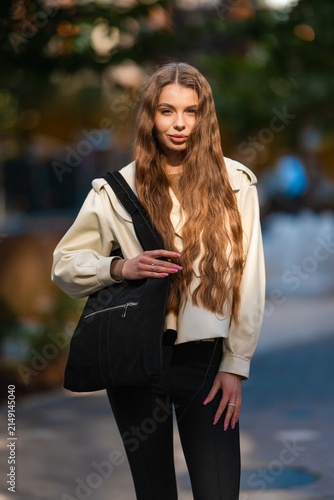 Young woman with long wavy brown hair wearing beige jacket and black tote bag outdoors in urban portrait with soft blurred background and gentle sunlight, with woman, female, young, adult detail