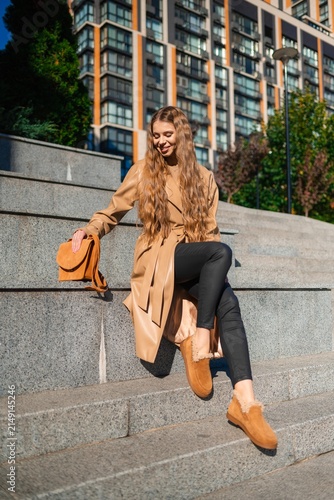 Young woman in tan trench coat sits on city steps beside orange handbag, framed by modern glass and brick architecture in daylight with relaxed smile and casual urban fashion style