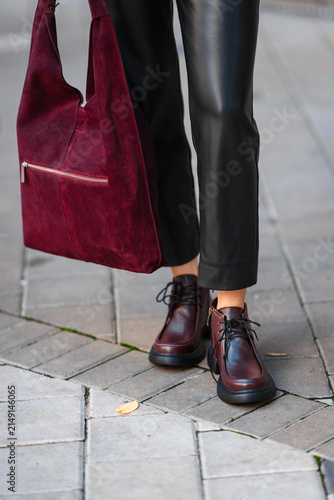 Burgundy suede handbag beside black leather pants and lace up boots on city sidewalk, creating modern urban fashion street style with rich textures and confident color contrast
