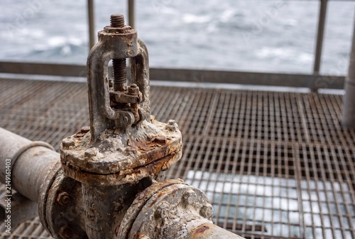 A close-up view of a heavily weathered and rusty industrial pipe valve on a metal grating floor of an offshore oil drilling platform.