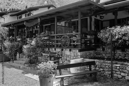 Black and white photograph of a traditional Italian osteria nestled in a mountain village, featuring rustic wooden architecture, stone walls, benches, wine barrels, and decorative plants. The peaceful