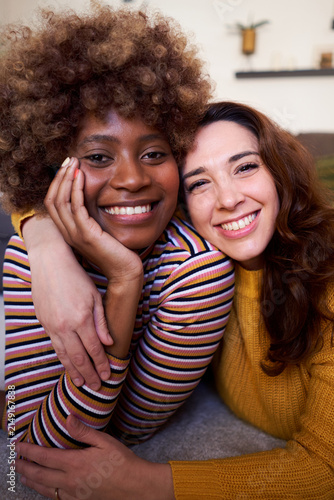 Cheerful portrait of two happy multi-ethnic women lying down in the living room, looking at camera smiling. Lesbian couple relaxing at home. Vertical image with copy space.