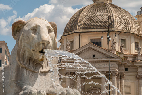 Lion fountain at Piazza del Popolo. Famous square in Rome, Italy at a sunny day with blue sky with Santa Maria in Montesanto in the background.