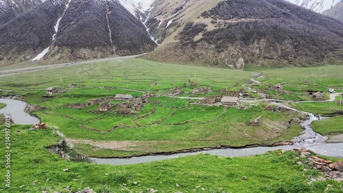Panoramic view of a remote mountain village with stone houses, winding river flowing through green pastures, snow-capped peaks in the background, tranquil rural landscape, Truso valley, Georgia