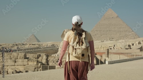Caucasian woman tourist walking towards Great Sphinx with ancient Egyptian pyramids at Giza Plateau in Cairo in background