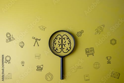STEM education and creative thinking concept. Magnifying glass focusing on a digital brain graphic surrounded by science and learning icons on a yellow background with copy space.