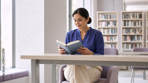 Woman reading a book at a desk in a library. Adult female student studying and researching in a modern academic environment