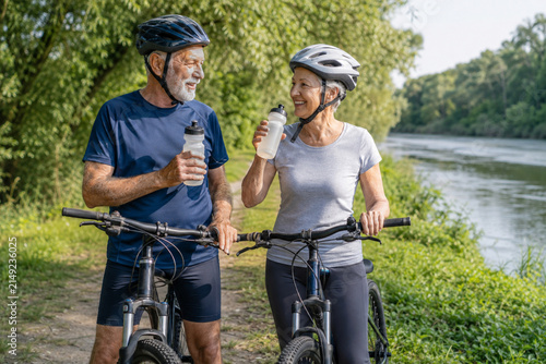 Senior Cyclists Hydrating by River: A happy elderly couple with bicycles pause their ride by a serene river, hydrating themselves while enjoying a sunny day in nature.