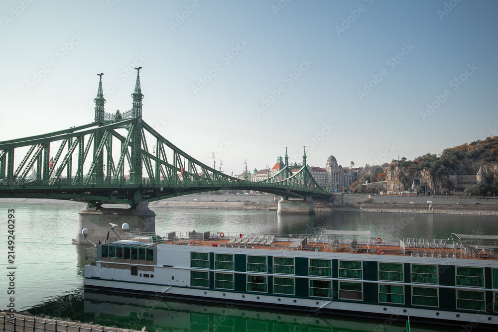Naklejka premium Liberty Bridge steel truss structure over the Danube River in Budapest, Hungary, with a river cruise ship docked below and Gell rt Hill visible in the background.