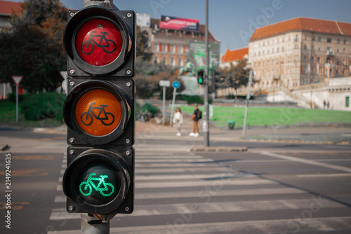 A green bicycle traffic light glows at a city crosswalk, with its red and amber signals above, set against a blurred European street scene with historic buildings and pedestrians.