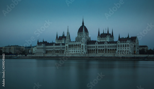 Photography The Hungarian Parliament Building's Gothic Revival spires and central dome rise along the calm Danube River at dusk, bathed in soft blue twilight tones in Budapest