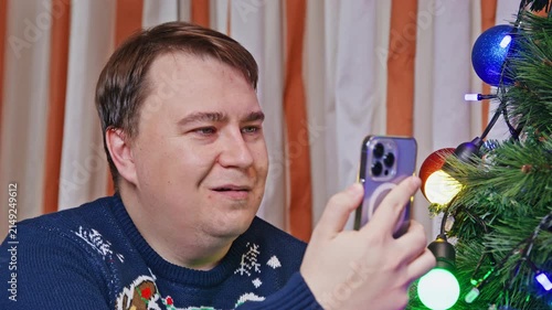 An adult man wearing a knitted Christmas sweater sits beside a decorated tree, raising a smartphone to take a photo and smiling as he checks the screen at home.
