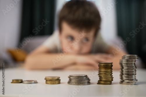 A blurry boy looks at stacks of coins on a table, close-up. Financial scarcity education. Children and pocket money. Savings, personal finance, modern education.