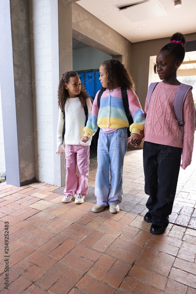 Fototapeta premium African American teenage girls holding hands and walking in school hall with backpacks near lockers
