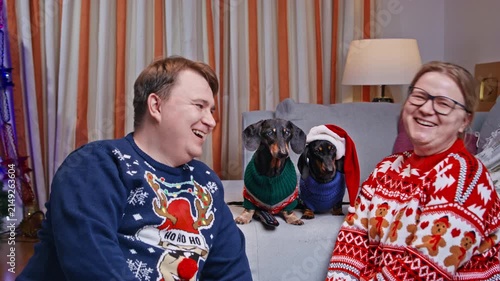 A man and woman in festive sweaters sit on a sofa laughing together, while two dachshund dogs dressed in tiny sweaters and a santa hat sit between them at home.