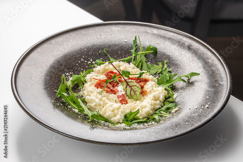 Creamy risotto served on a black plate garnished with arugula, sun-dried tomatoes, and herbs on a white table in a modern dining setting