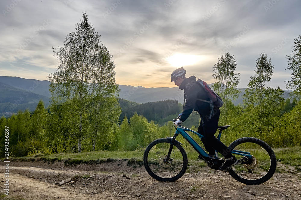 Fototapeta premium Side view of mountain biker riding electric bike on dirt trail through green forest hills at sunset, scenic landscape and outdoor adventure concept