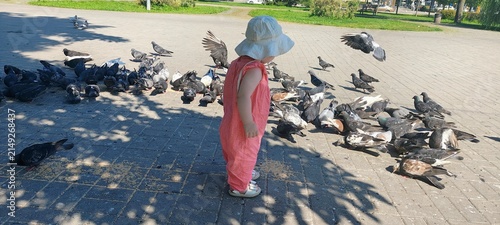Adorable toddler girl in sun hat standing near pigeons on paved square in summer