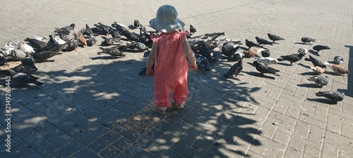 Adorable toddler girl in sun hat standing near pigeons on paved square in summer
