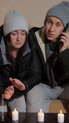 Worried couple wearing warm hats and jackets trying to stay warm over candle flames during a blackout, as the anxious man calls for help with the broken central heating system