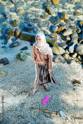 Photography High-angle full-length shot of a young Asian Muslim woman in a cream hijab, long-sleeved shirt, and pleated skirt standing on a rocky sandy beach