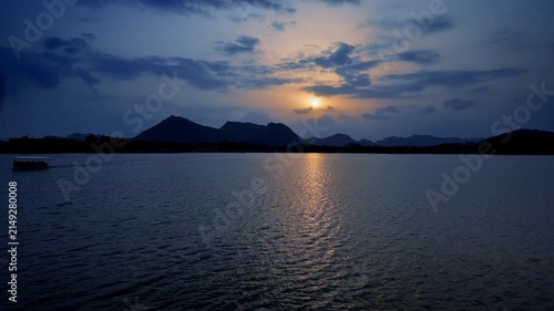 Golden light streams across rippling water as dusk falls over distant peaks. A small boat moves along the quiet horizon. Evening skies glow above Udaipur�s tranquil lake.