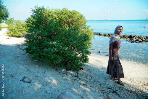 Fotografie ​A cheerful young Asian Muslim woman wearing a hijab, long-sleeved top, and pleated skirt smiling as she walks along a sandy tropical beach near coastal bushes