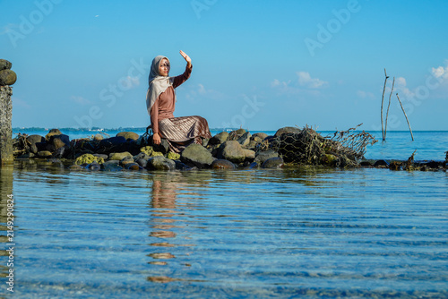 Obraz na płótnie ​A young Asian Muslim woman in a hijab and stylish pleated skirt sitting on coastal rocks, raising her hand to shield her eyes from intense sunlight