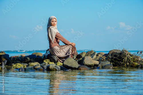 Full-length portrait of a young, elegant South East Asian Muslim woman wearing a beige hijab, brown long-sleeve shirt, and a stylish pleated skirt. She is sitting gracefully on a natural barrier of ro